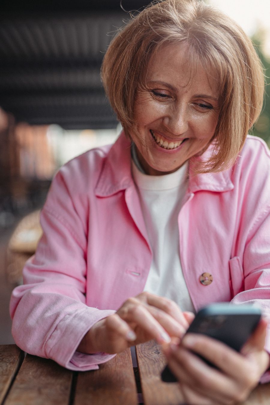 A woman in her sixties at an outdoor cafe table, smiling as she goes through her phone