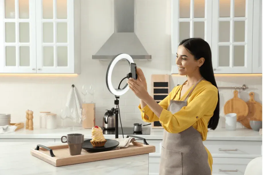 A woman filming herself in her kitchen with a smartphone on a ring light